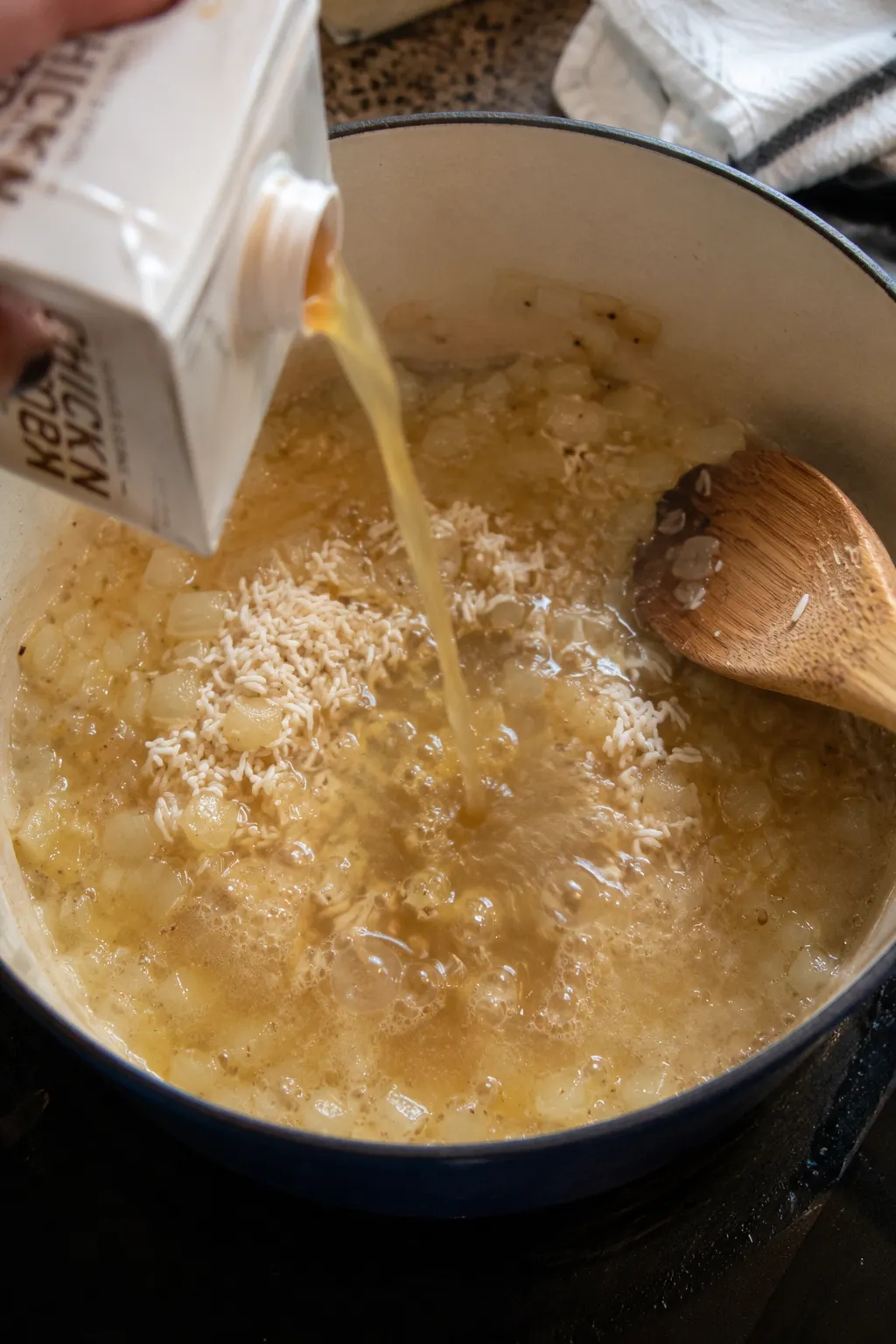 Rice being stirred into a pot with sauteed onion and chicken broth being poured in