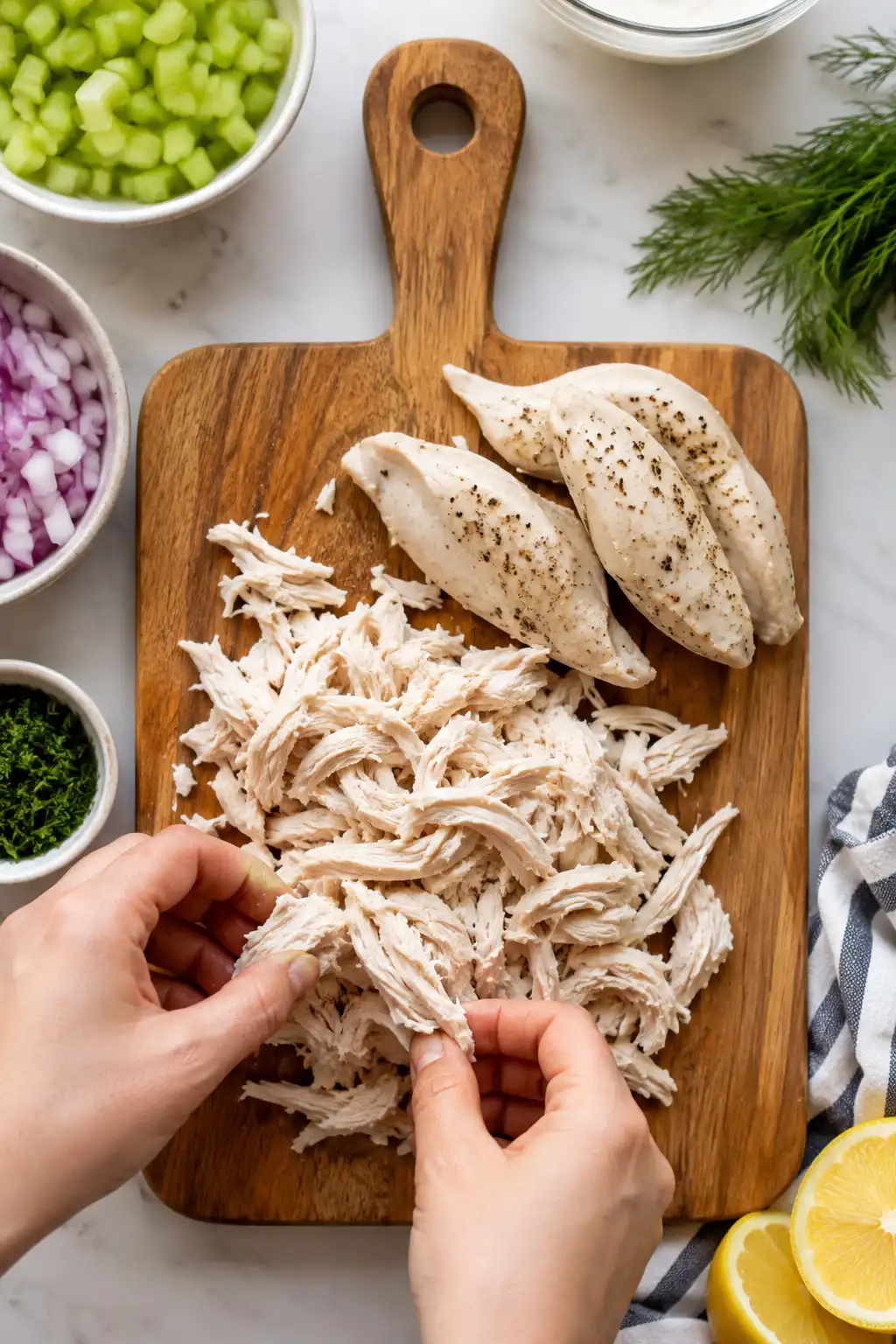 Hands shredding cooked chicken breast into bite sized pieces on a cutting board