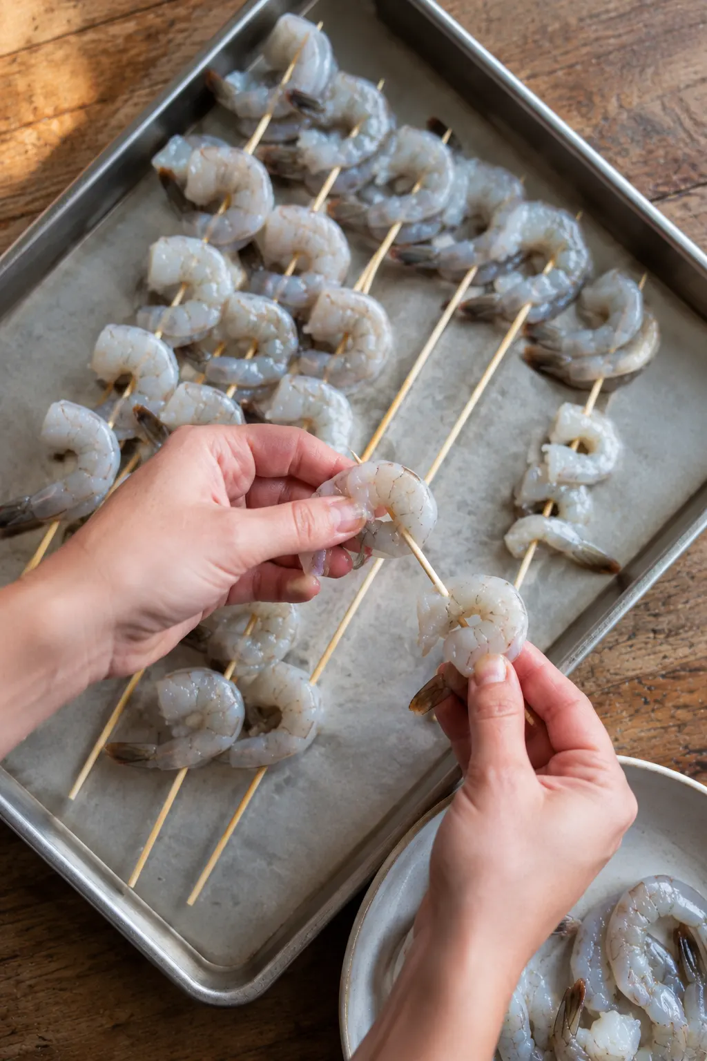 Raw shrimp being threaded onto wooden skewers on a baking sheet
